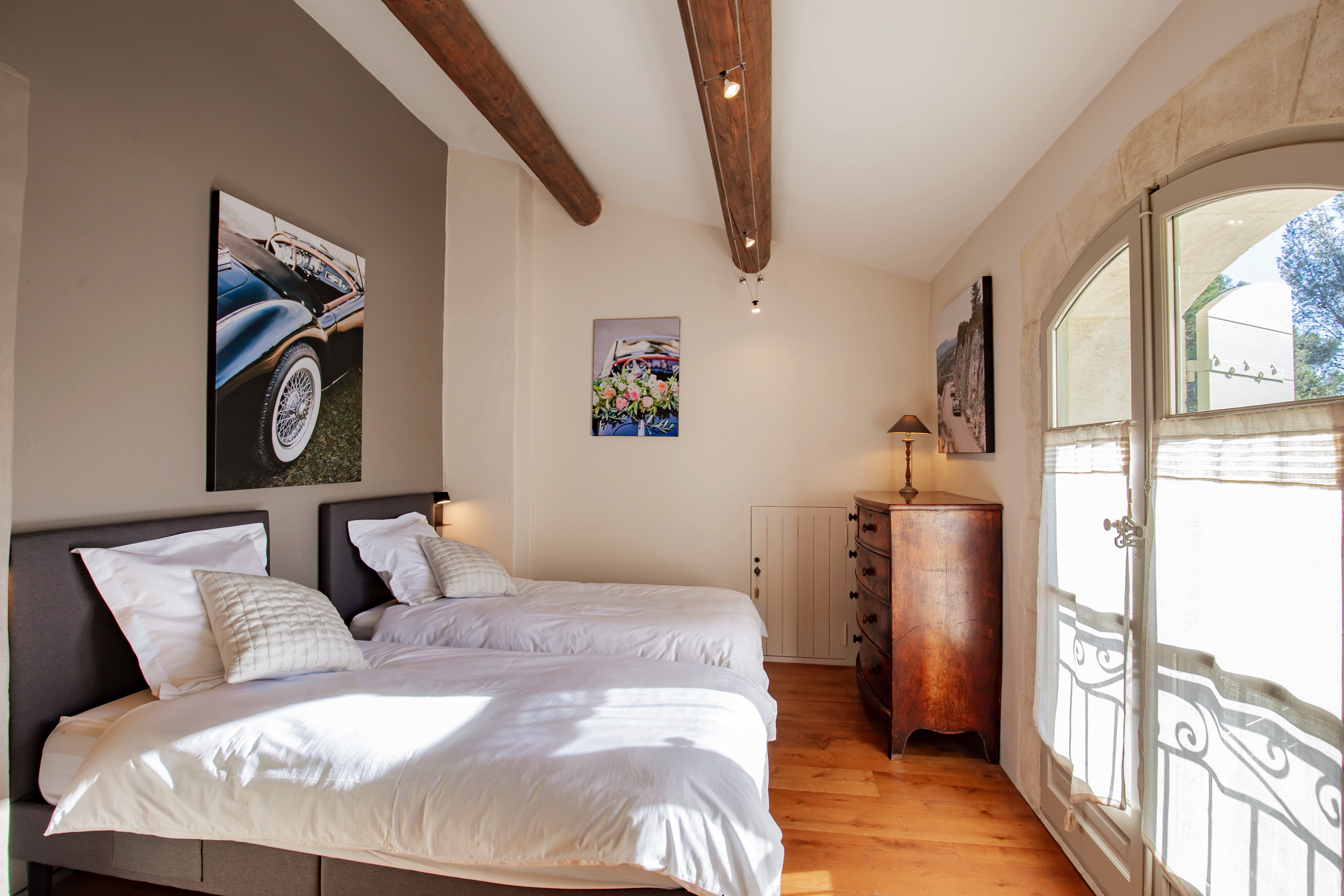 A bright, cozy twin bedroom featuring wooden beams, white linens, framed artwork above the beds, a vintage wooden dresser, and a bedside lamp. A window lets in natural light.